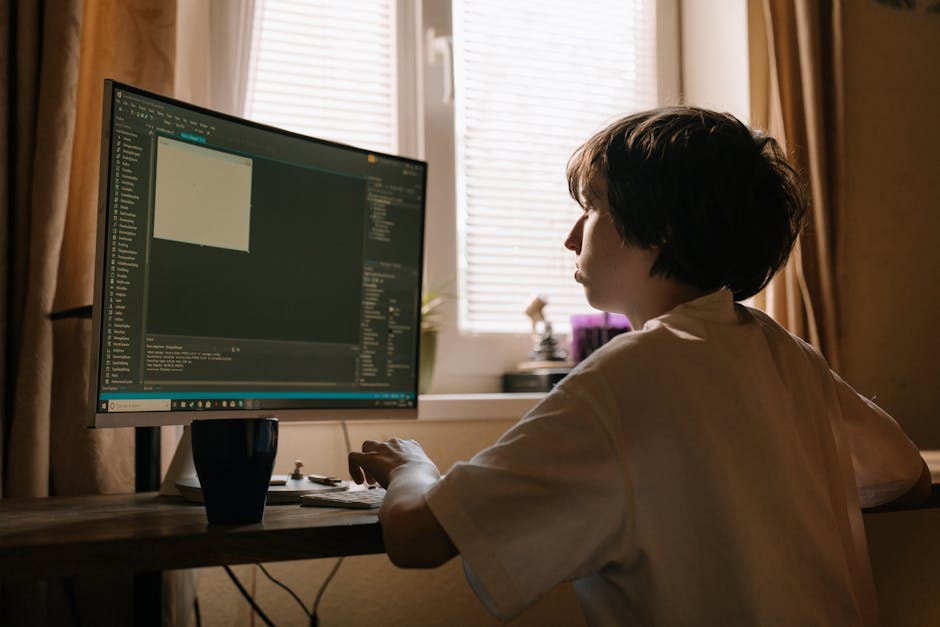 A person with short dark hair sitting at a wooden desk indoors, focused on a large desktop computer with a dark-themed coding interface on the screen. The background features a window with horizontal 