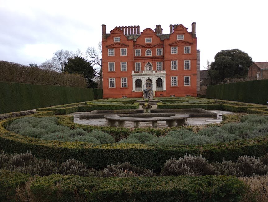 The image depicts a historic red-brick mansion with a symmetrical faÃ§ade, multiple small windows, and decorative architectural details, set against an overcast sky. In the foreground, a formal garden